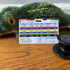May include: A green plush alligator toy rests near a reference card with medical information for pediatric patients. A black stethoscope is also in the image. The card includes age ranges and vital signs.