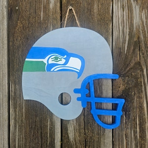 May include: A wooden, gray football helmet with a blue facemask and a logo of a blue and white bird with green accents. The helmet is hanging by a natural twine string against a weathered wood background.