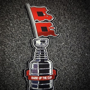 May include: A sticker featuring a black and white illustration of a trophy cup with a red banner across the front that reads RAISE UP THE CUP. Above the cup, two red flags with black square centers are attached to a single flagpole.