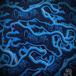 May include: A collection of stickers featuring a black panther design with the word "CAROLINA" in a stylized font. The panther is outlined in light blue, with a dark blue background. The stickers are layered on top of each other.