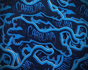 Op de afbeelding: Een verzameling stickers met een zwart panterontwerp en het woord "CAROLINA" in een gestileerd lettertype. De panter is omlijnd in lichtblauw, met een donkerblauwe achtergrond. De stickers zijn op elkaar gelegd.