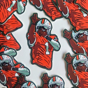 May include: A collection of stickers featuring an American football player in an orange jersey with the number 1. The player is wearing a white helmet with a green and orange stripe. The stickers have a dark green border.