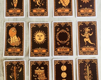 May include: A set of 12 tarot cards with gold trim and black backgrounds. The cards feature illustrations of various figures and symbols, including a lion, a sun, a moon, a sword, a cup, a wand, a star, a fool, a devil, death, justice, and the ace of rings.
