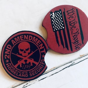 May include: Two round, red and black coasters. One coaster features a skull and crossbones with the text "2nd Amendment", "America's Original Homeland Security", and "1789". The other coaster features a distressed American flag with the text "We The People".