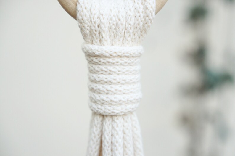 May include: Close-up of a white macrame plant hanger with a wooden ring at the top. The macrame is made with thick cotton cord and has a knot at the bottom.