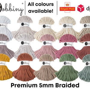 May include: Assortment of 15 spools of 5mm braided cotton rope in various colours, including white, beige, grey, pink, red, yellow, and green. The spools are arranged in three rows of five. The text "Bobbiny" and "Premium 5mm Braided" are visible at the top and bottom of the image.