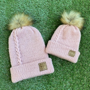 May include: Two pink knitted hats with faux fur pom poms. The larger hat has a brown leather patch with the word "MAMA" on it. The smaller hat has a brown leather patch with the word "MINI" on it.