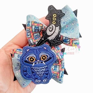 May include: A hair bow featuring a blue glittery fabric base with a pattern of framed blue cat illustrations. It is adorned with two embroidered patches: a black and white stylized creature and a blue cat with large orange eyes and sharp teeth.