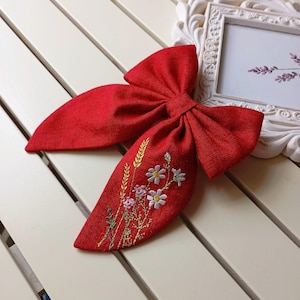 Könnte beinhalten: Eine leuchtend rote Haarspange aus Leinen mit gestickten floralen Details. Die Schleife hat einen zentralen Knoten und lange, fließende Enden. Die Stickerei umfasst Weizen und zarte Blumen in Weiß, Rosa und Gelb.