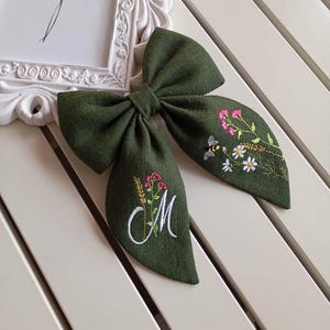 Op de afbeelding: Een donkergroene stoffen strik met geborduurde bloemen en bijen details. De strik heeft een wit geborduurde letter "M" en delicate bloemmotieven in roze, geel en wit. Een haaraccessoire.