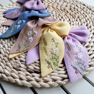 Pode incluir: Cinco laços de cabelo de tecido com desenhos florais bordados. Os laços são em tons de rosa, azul, marrom, amarelo e roxo. Os laços estão dispostos sobre um tapete de palha trançado.