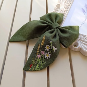 Könnte beinhalten: Eine grüne Stoffschleife mit gestickten Wildblumen und Weizenähren. Die Schleife ist zu einem Knoten gebunden und hat einen rustikalen, natürlichen Look.