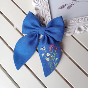 May include: A blue fabric bow with an embroidered design of flowers and a bee. The bow is tied with a knot in the centre.