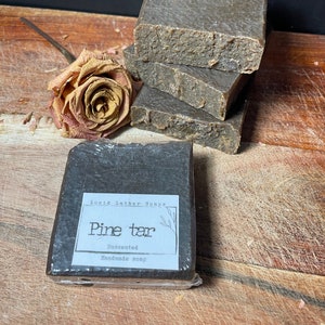 May include: A bar of dark brown pine tar soap wrapped in plastic with a white label that reads "Lucid Lather Soaps Pine tar Unscented Handmade soap".