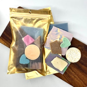 May include: A gold zippered pouch filled with various colorful handmade soap bars. The soap bars are in different shapes and sizes, including hearts, circles, and squares. The soap bars are arranged on a wooden surface.