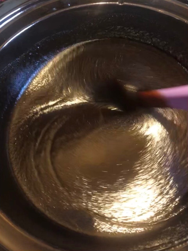 May include: A close-up of a dark brown liquid being stirred in a silver pot with a pink spatula.