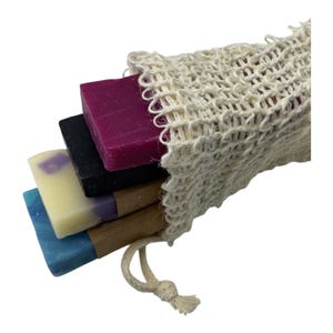 May include: A collection of colorful handmade soaps in a natural mesh bag. The soaps are various colors, including purple, black, yellow, and blue. The bag is made of a woven, off-white material and has a drawstring closure.