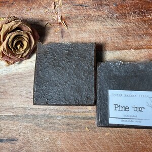 May include: Two bars of dark brown soap on a wooden surface. The soap on the right has a white label that reads "Lucid Leather Soaps Pine tar Unscented Handmade Soap".