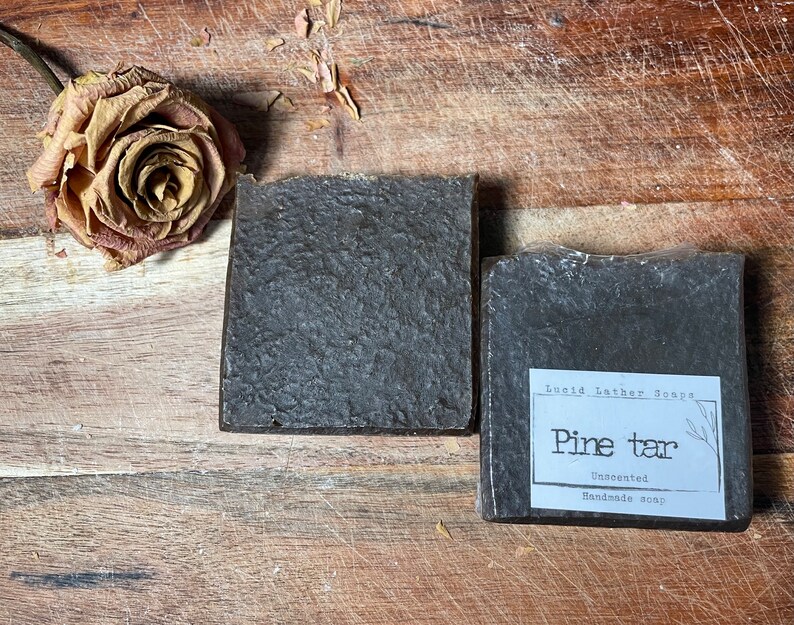 May include: Two bars of dark brown soap on a wooden surface. The soap on the right has a white label that reads "Lucid Leather Soaps Pine tar Unscented Handmade Soap".