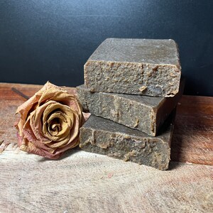May include: Three bars of brown soap stacked on top of each other on a wooden surface. A dried rose is next to the soap.