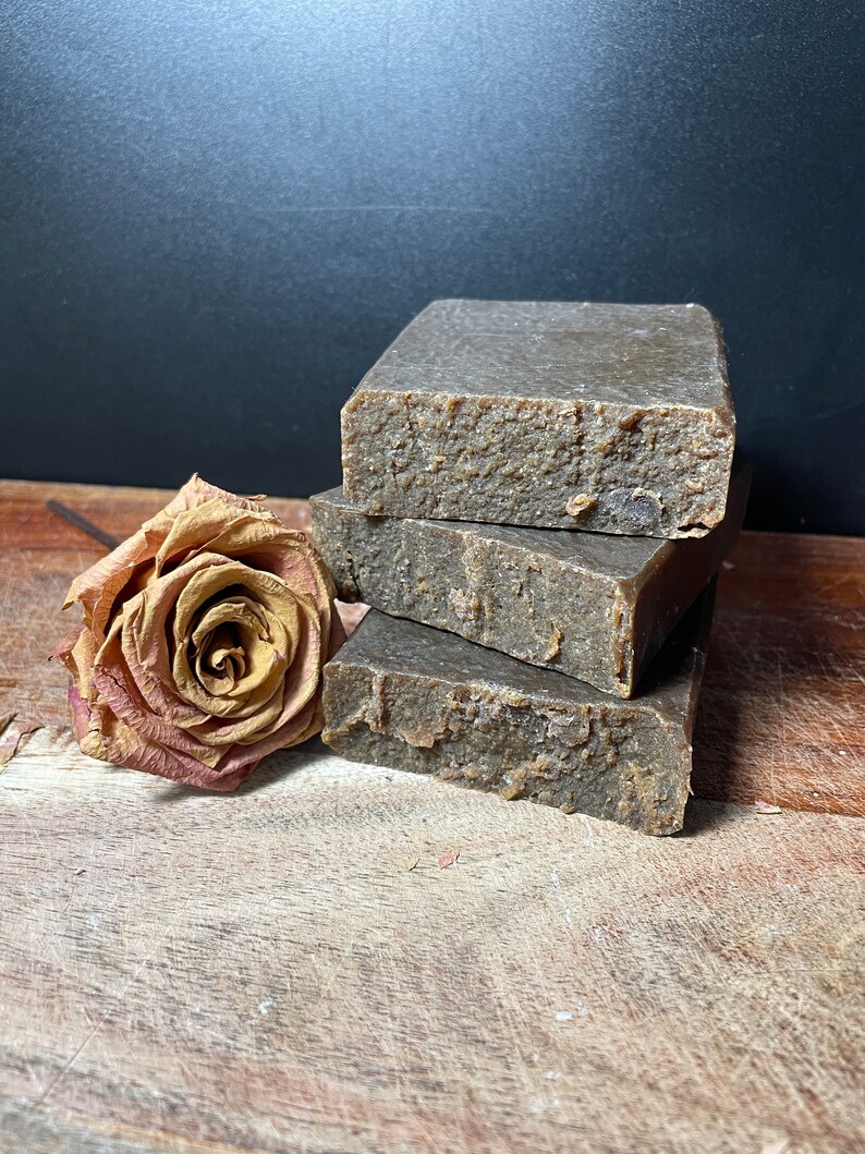 May include: Three bars of brown soap stacked on top of each other on a wooden surface. A dried rose is next to the soap.