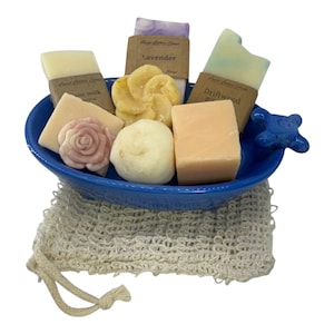 May include: A collection of assorted soap bars and shapes in a blue ceramic dish with a faucet handle detail. The set includes rectangular bars with paper labels, a rose-shaped soap, and round soaps, all resting on a beige textured mesh soap bag.