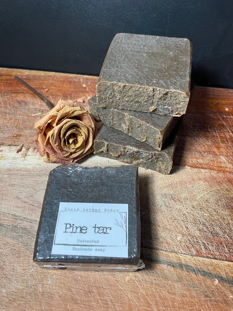May include: A bar of dark brown pine tar soap wrapped in plastic with a white label that reads "Lucid Lather Soaps Pine tar Unscented Handmade soap".