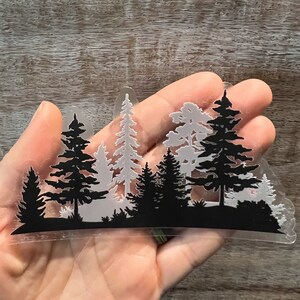 May include: A clear sticker featuring a black silhouette of a forest scene. The sticker depicts multiple pine trees of varying sizes, with some trees in the background appearing lighter in colour.