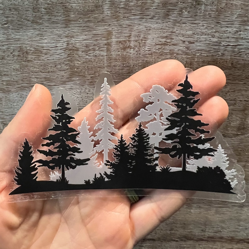 May include: A clear sticker featuring a black silhouette of a forest scene. The sticker depicts multiple pine trees of varying sizes, with some trees in the background appearing lighter in colour.