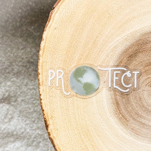 May include: A white sticker with the word "PROTECT" in a stylized font with a green and white globe in the center of the word. The sticker is on a light brown wooden surface.