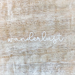 May include: White cursive lettering on a light brown wood background that reads "wanderlust".