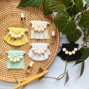 May include: Five macrame wall hangings with wooden beads, each in a different color: yellow, white, green, black, and white. The hangings are displayed on a woven wicker tray with small glass bottles.