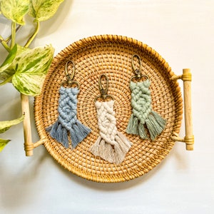 May include: Three handmade macrame keychains with tassels in a light blue, cream, and green color scheme. The keychains are displayed on a woven tray.