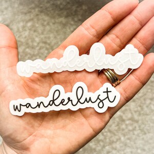 May include: Two white stickers with the word "wanderlust" written in black script. The top sticker has the word in white script.
