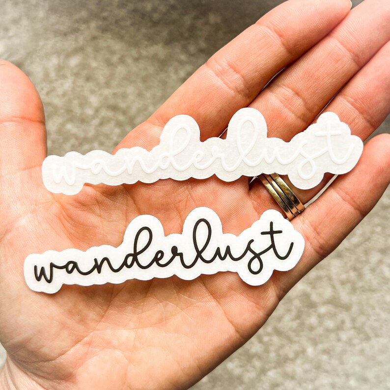 May include: Two white stickers with the word "wanderlust" written in black script. The top sticker has the word in white script.