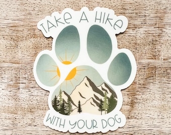 May include: A white paw print sticker with a mountain scene and a sun inside. The sticker says "Take a hike with your dog" in a blue and white font.