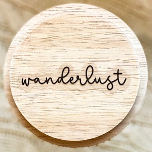 May include: A round wooden coaster with the word "wanderlust" written in black cursive lettering.