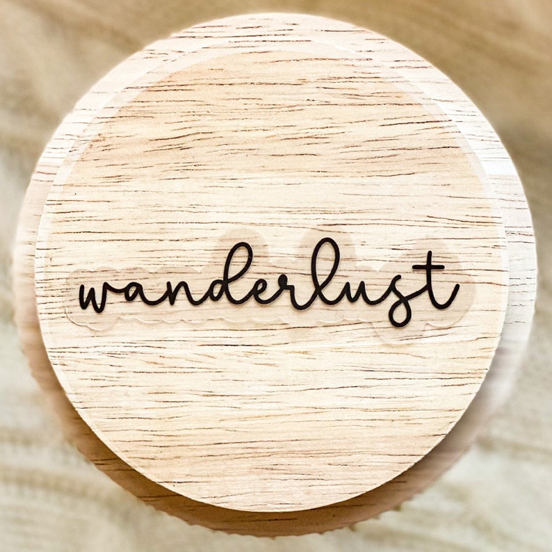May include: A round wooden coaster with the word "wanderlust" written in black cursive lettering.