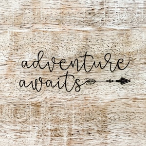 以下が含まれることがあります： 黒い筆記体で「adventure awaits」と書かれた透明なステッカー。矢印と羽と矢じりのデザインがテキストの下にあります。ステッカーは、明るい茶色の質感のある木製の表面にあります。