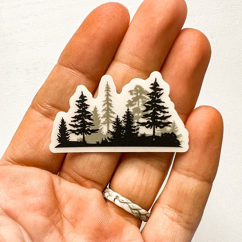 May include: A die-cut sticker featuring a forest silhouette in shades of black, gray, and white. The design includes various tree types, creating a layered effect. The sticker is held in a hand.