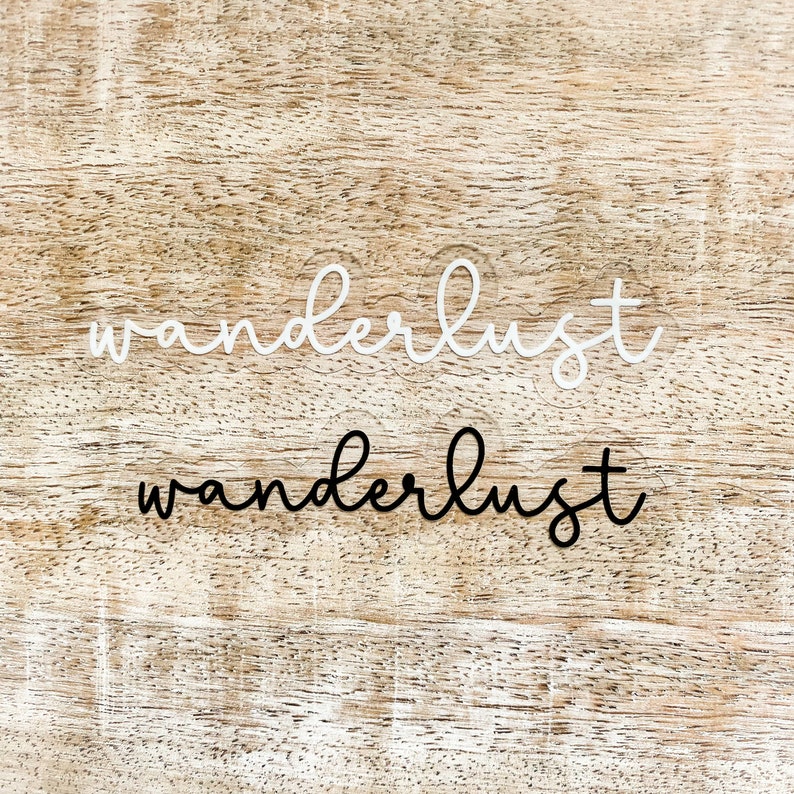 May include: Two versions of the word "wanderlust" written in cursive, one in white and one in black, on a light brown wooden background.