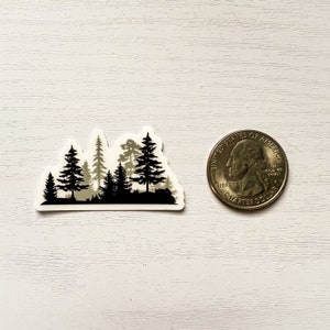 May include: A sticker featuring a forest silhouette in black and gray tones. The design includes various tree shapes and a dark base, with a quarter coin for size comparison. The sticker is on a white surface.