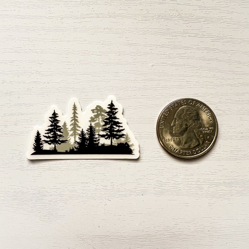 May include: A sticker featuring a forest silhouette in black and gray tones. The design includes various tree shapes and a dark base, with a quarter coin for size comparison. The sticker is on a white surface.