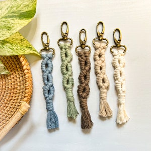 May include: Five colorful macrame keychains with gold key rings. The keychains are made with different colored cords and have a tassel at the bottom. The keychains are arranged in a row on a white surface.