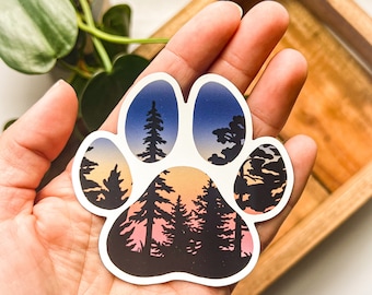 May include: A paw print sticker with a forest scene inside. The paw print is white with a black outline. The forest scene is a sunset with trees and a blue sky.