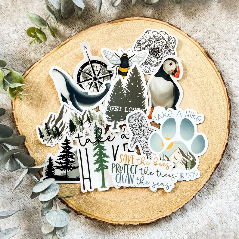 May include: A collection of colorful stickers with nature-themed designs. The stickers feature illustrations of trees, a compass, a bee, a puffin, a paw print, and text that reads "Save the bees, protect the trees, clean the seas".