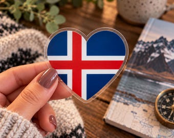 May include: A heart-shaped sticker featuring the national flag of Iceland, which consists of a blue background with a red cross outlined in white. The sticker has a clear border and is held up against a blurred background of a book and greenery.