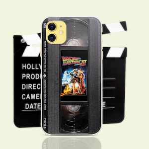 Puede incluir: Una funda para iPhone negra y plateada con un diseño de cinta VHS vintage. La funda presenta un título de película "Regreso al futuro III" y una claqueta negra y blanca.