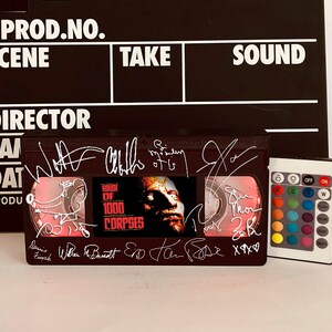 Pode incluir: Uma claquete preta e branca com as palavras "PROD.NO., SCENE, TAKE, SOUND, DIRECTOR, CAM, DAT, PRODU" impressas nela. Uma fita VHS com o título "HOUSE OF 1000 CORPSE" está abaixo da claquete. A fita tem várias assinaturas. Um controle remoto com botões coloridos está à direita da fita.