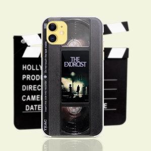 May include: A black and silver phone case with a vintage VHS tape design. The case features the title "The Exorcist" and has a black and white image of two figures walking down a street. The case has a clear plastic window on the back.